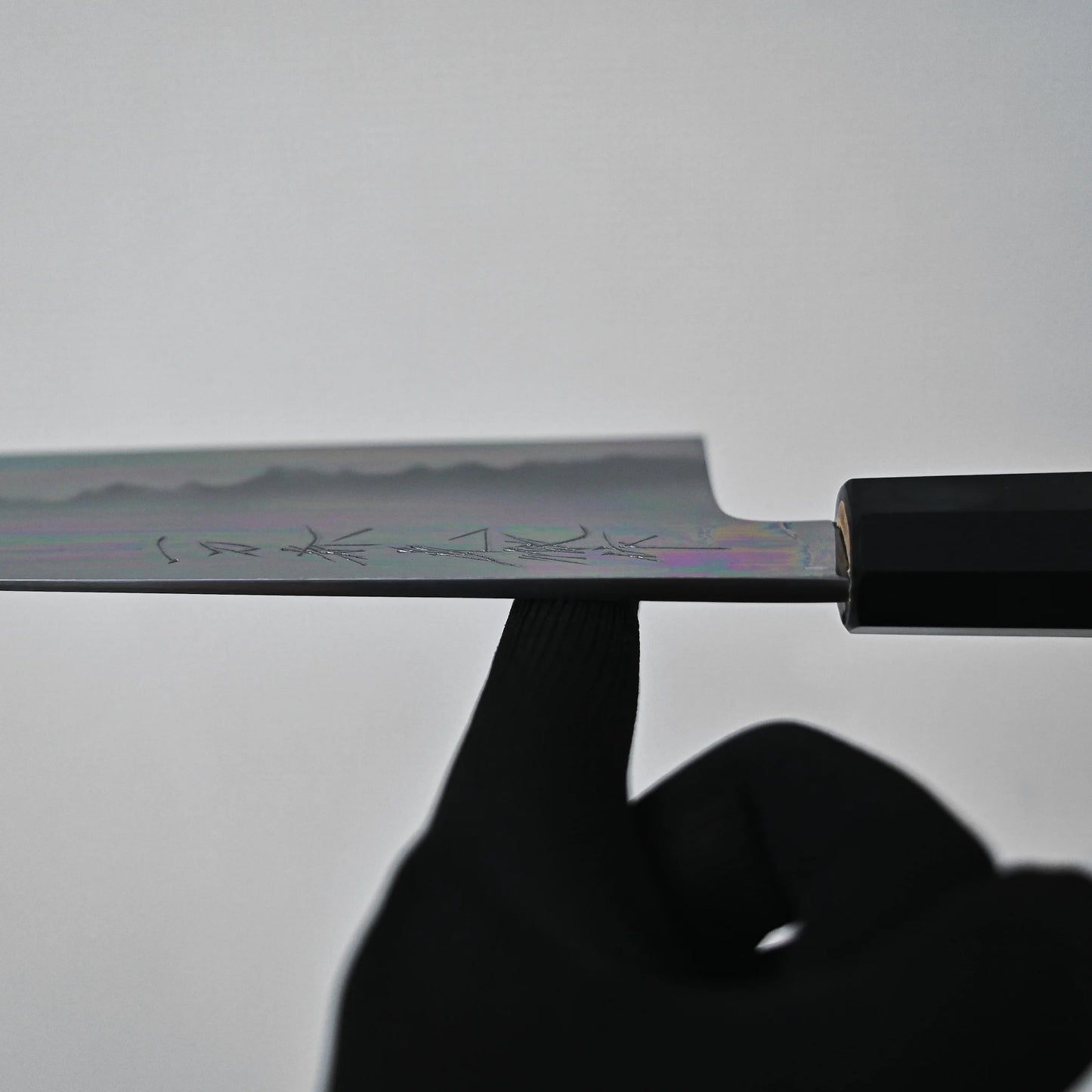 A black-gloved hand balances the Sakai knife by its blade on the tip of an index finger. Faint engravings and the Aogami#1 core are visible against a plain, light-colored background.