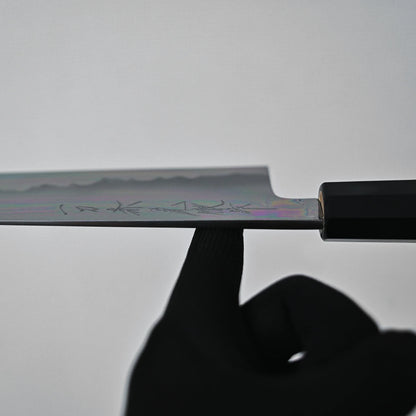 A black-gloved hand balances the Sakai knife by its blade on the tip of an index finger. Faint engravings and the Aogami#1 core are visible against a plain, light-colored background.