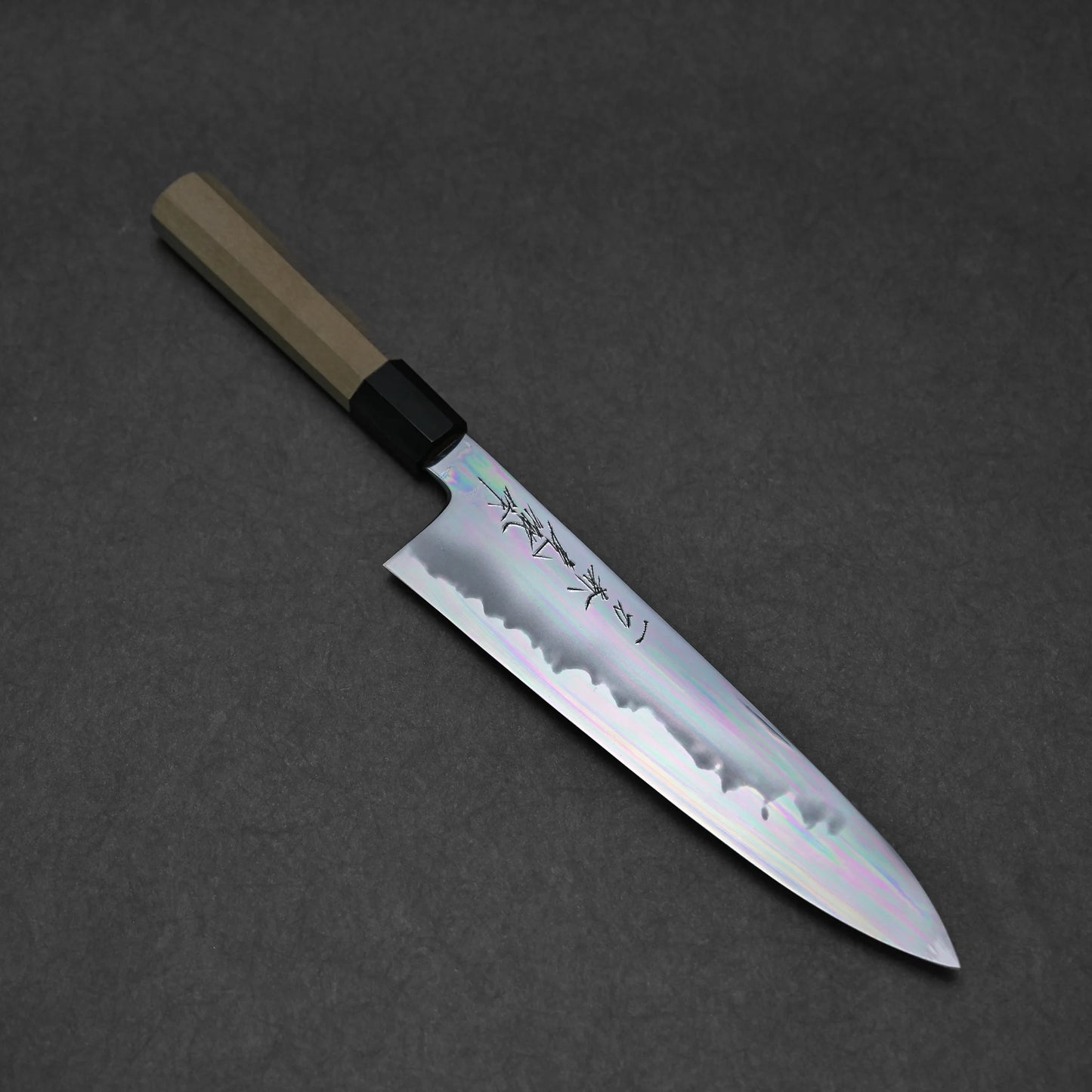 A tall gyuto features a shiny, engraved silver blade with an aogami#1 core and wooden handle, shown on a dark gray surface.