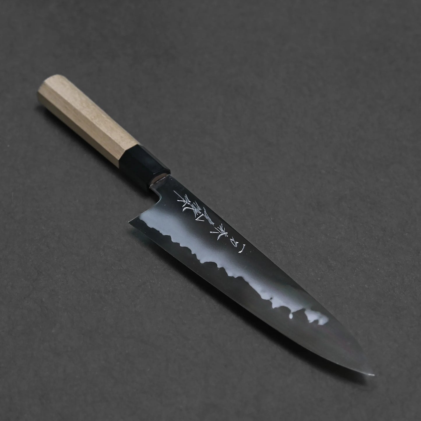 The carbon gyuto features a light wooden handle and a shiny black blade engraved with Japanese characters, beautifully displayed on a dark background.