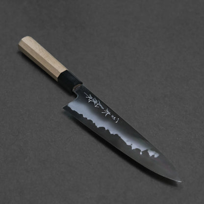 The carbon gyuto features a light wooden handle and a shiny black blade engraved with Japanese characters, beautifully displayed on a dark background.