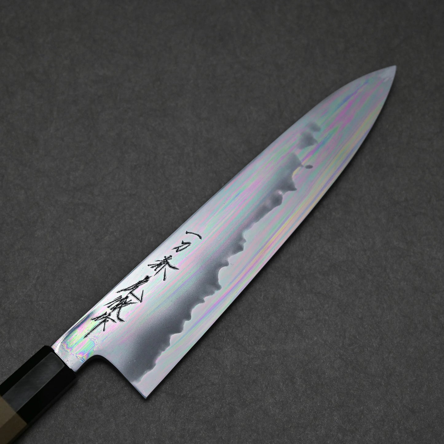 A close-up of a tall gyuto, showing its polished blade with wavy hamon and engraved kanji, set against a dark textured background.
