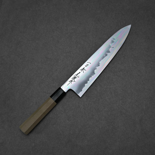 The Yoshikazu Tanaka aogami#1 tall gyuto 230x54mm, featuring a wooden handle and patterned steel blade engraved with Japanese characters, rests on a dark gray textured surface.