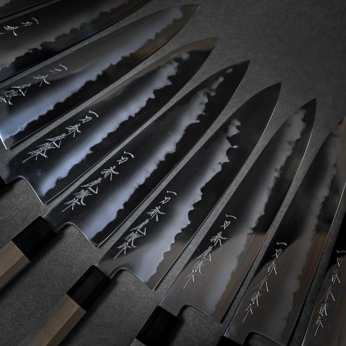A close-up shows six Japanese chef's knives with shiny engraved blades all arranged in parallel on a dark surface.