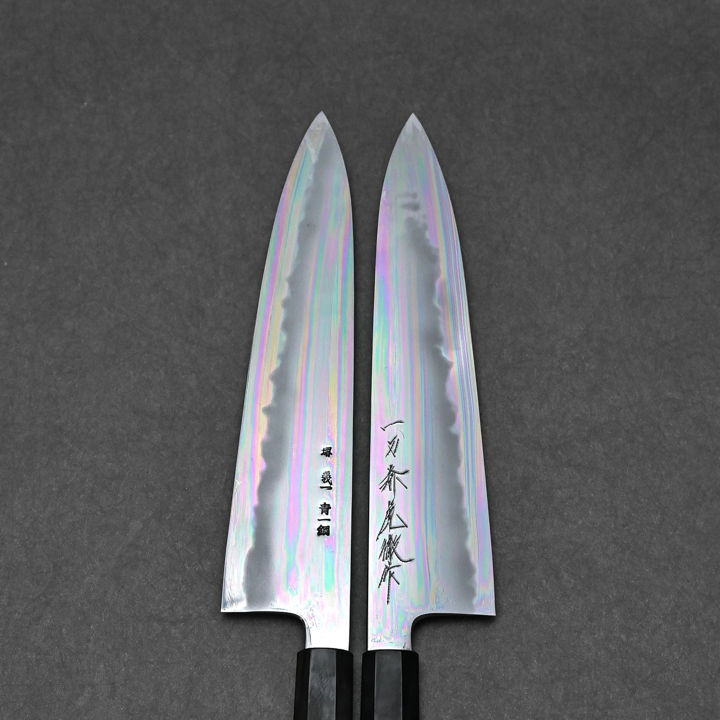 Two Tanaka knives, each with iridescent, polished blades engraved with unique Japanese characters, are side by side on a dark gray textured background, showcasing Tanaka’s exceptional craftsmanship.