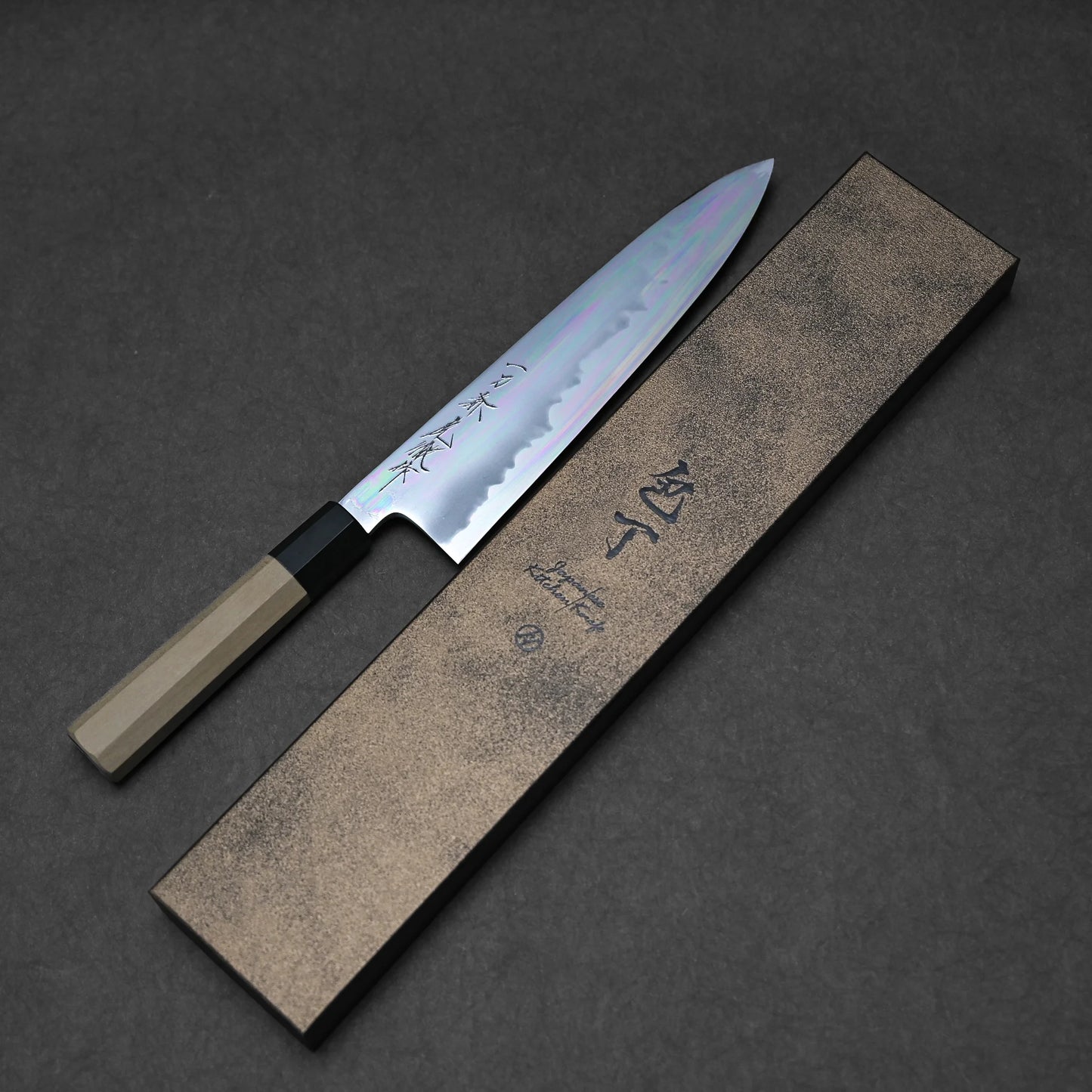 The Sakai aogami#1 gyuto, featuring a wooden handle and crafted in Japan, sits beside a rectangular brown box with black Japanese characters on the lid, shown on a dark gray surface.