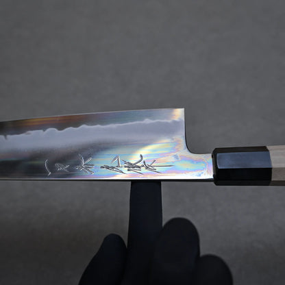 A hand in a black glove shows the balance point of a Japanese chef knife. Japanese characters are engraved on the shiny blade of a tall gyuto knife with a wooden handle, set against a plain gray background.