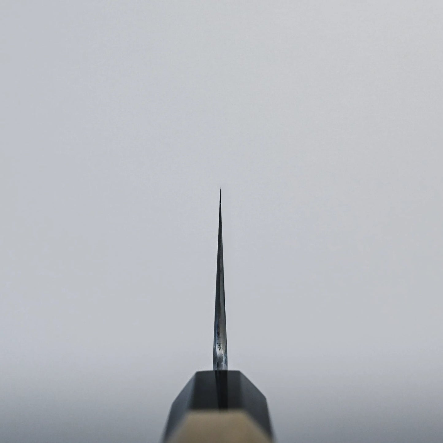 Choil shot of a Japanese chef's knife against a plain, light grey background. The blade is centered in the image and points vertically upward.