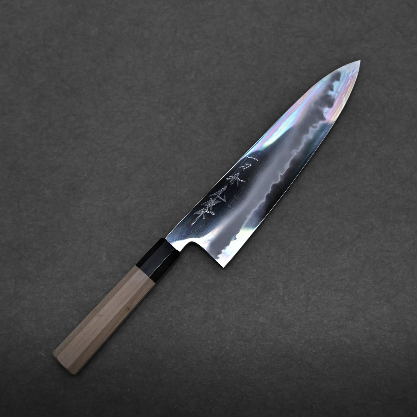 A Yoshikazu Tanaka aogami#1 extra tall gyuto 240mm with a wooden handle and a shiny, reflective blade featuring engraved characters rests on a dark, textured surface.