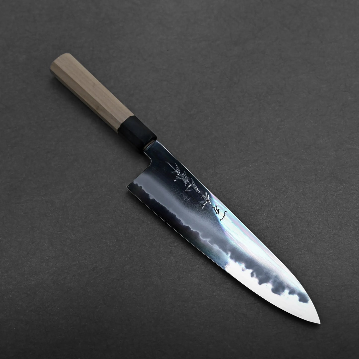 A tall gyuto knife with a wooden handle and shiny blade featuring engraved characters, lying on a dark, textured surface.