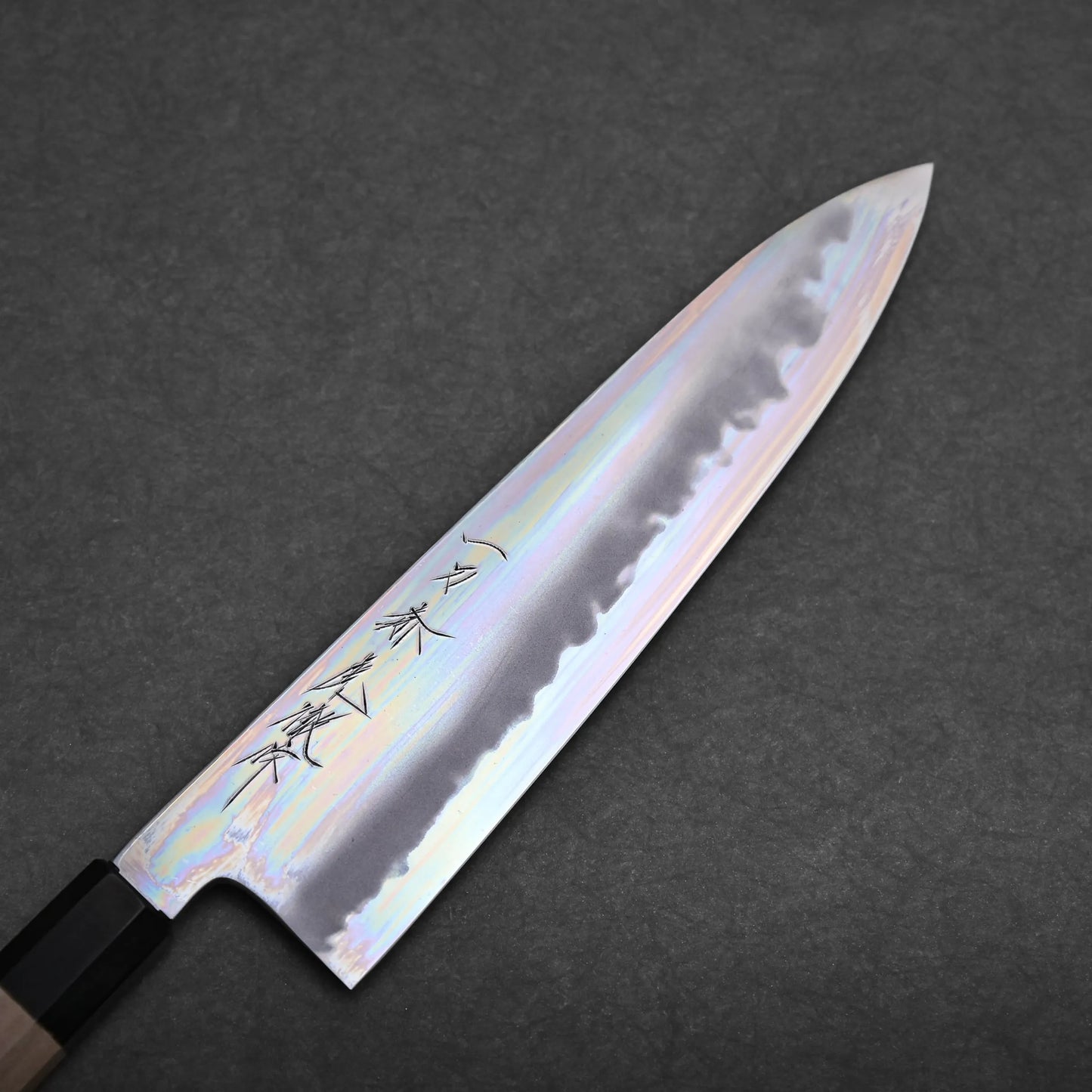 A tall gyuto knife with a shiny, iridescent blade and black handle rests on a dark, textured surface. The blade features engraved Japanese characters near the handle.