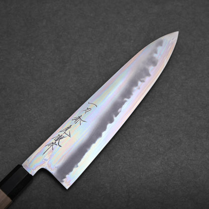 A tall gyuto knife with a shiny, iridescent blade and black handle rests on a dark, textured surface. The blade features engraved Japanese characters near the handle.