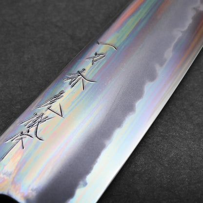 A close-up of a polished tall gyuto knife blade featuring engraved Japanese characters and a distinct, wavy hamon line, with iridescent reflections on the metal surface. The background is a dark, textured material.