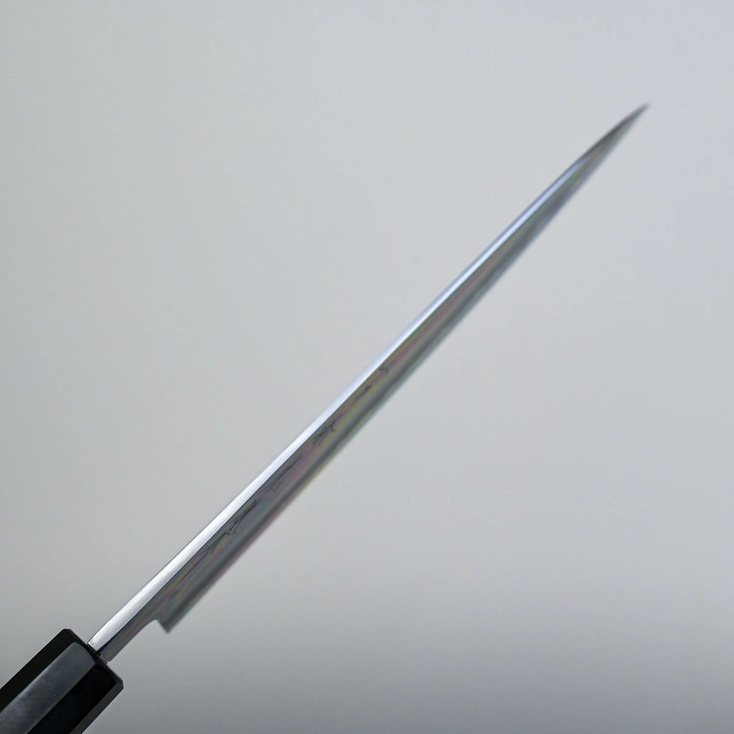 A spine shot of a tall gyuto knife blade is shown from the side, with the sharp edge facing forward and the handle partially visible in the lower left corner against a plain, light background.