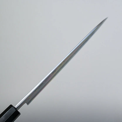 A spine shot of a tall gyuto knife blade is shown from the side, with the sharp edge facing forward and the handle partially visible in the lower left corner against a plain, light background.