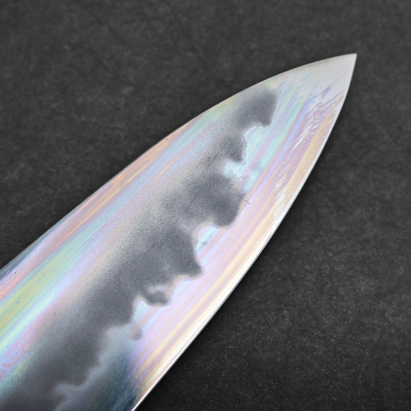 Close-up of a tall gyuto knife blade showing a wavy hamon pattern and an iridescent, rainbow-like finish, set against a dark, textured background.