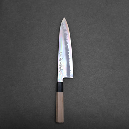 A tall gyuto knife with a light wooden handle and a shiny, reflective blade featuring engraved Japanese characters, placed on a dark, textured surface.
