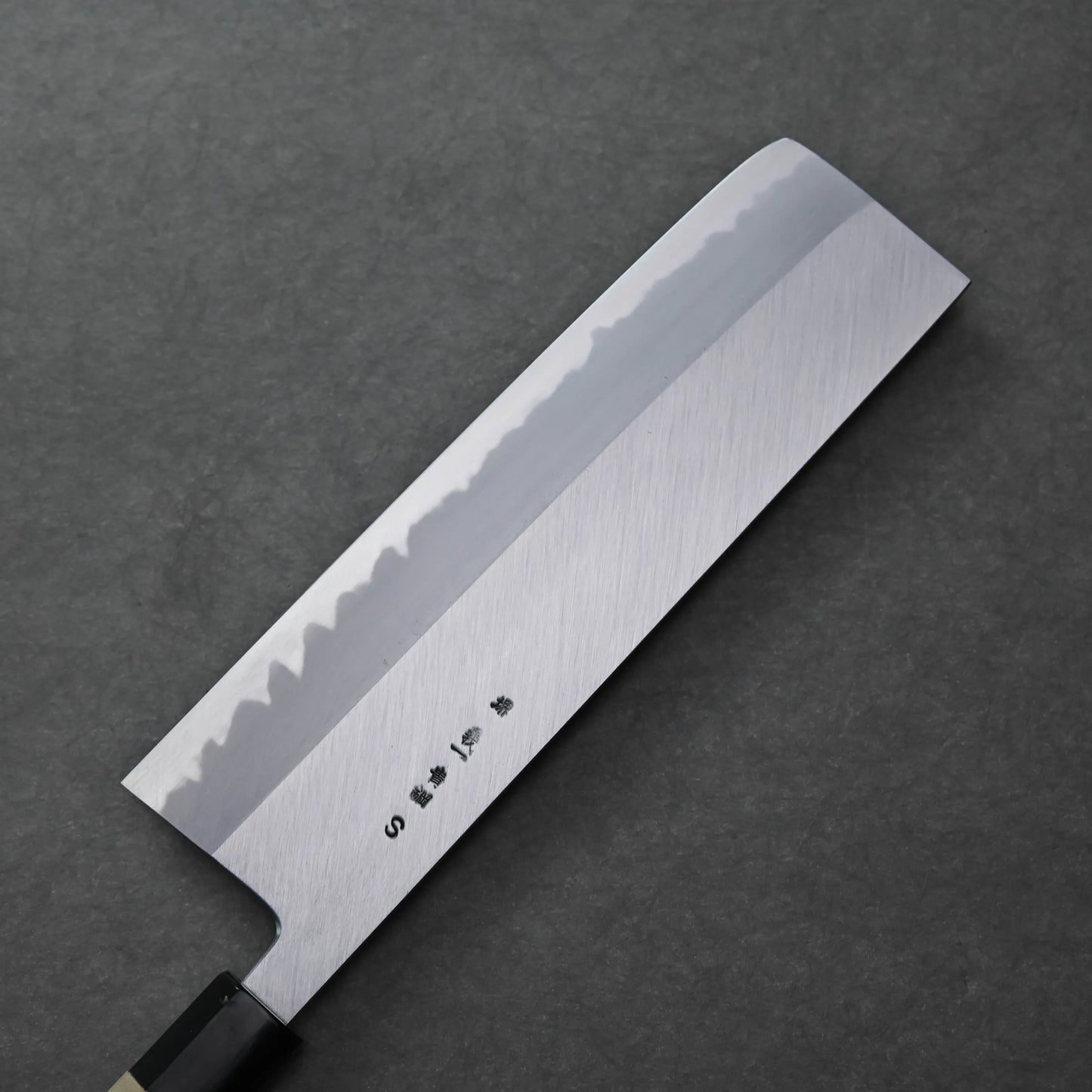 A Tanaka 210mm nakiri Japanese-style knife with a rectangular blade, light wavy pattern near the edge, and Japanese characters engraved on the side, placed on a dark textured surface.