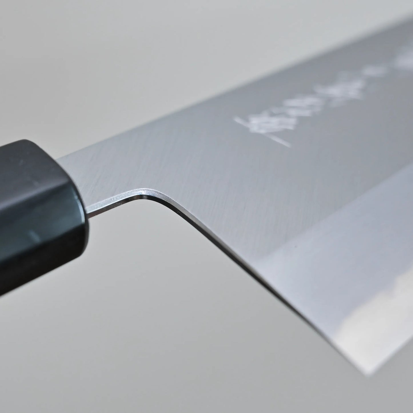 Close-up of the choil finish of a Tanaka 210mm nakiri chef’s knife, showing the brushed metal surface. Some engraved text is faintly visible on the blade. The background is plain and out of focus.