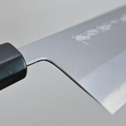 Close-up of the choil finish of a Tanaka 210mm nakiri chef’s knife, showing the brushed metal surface. Some engraved text is faintly visible on the blade. The background is plain and out of focus.