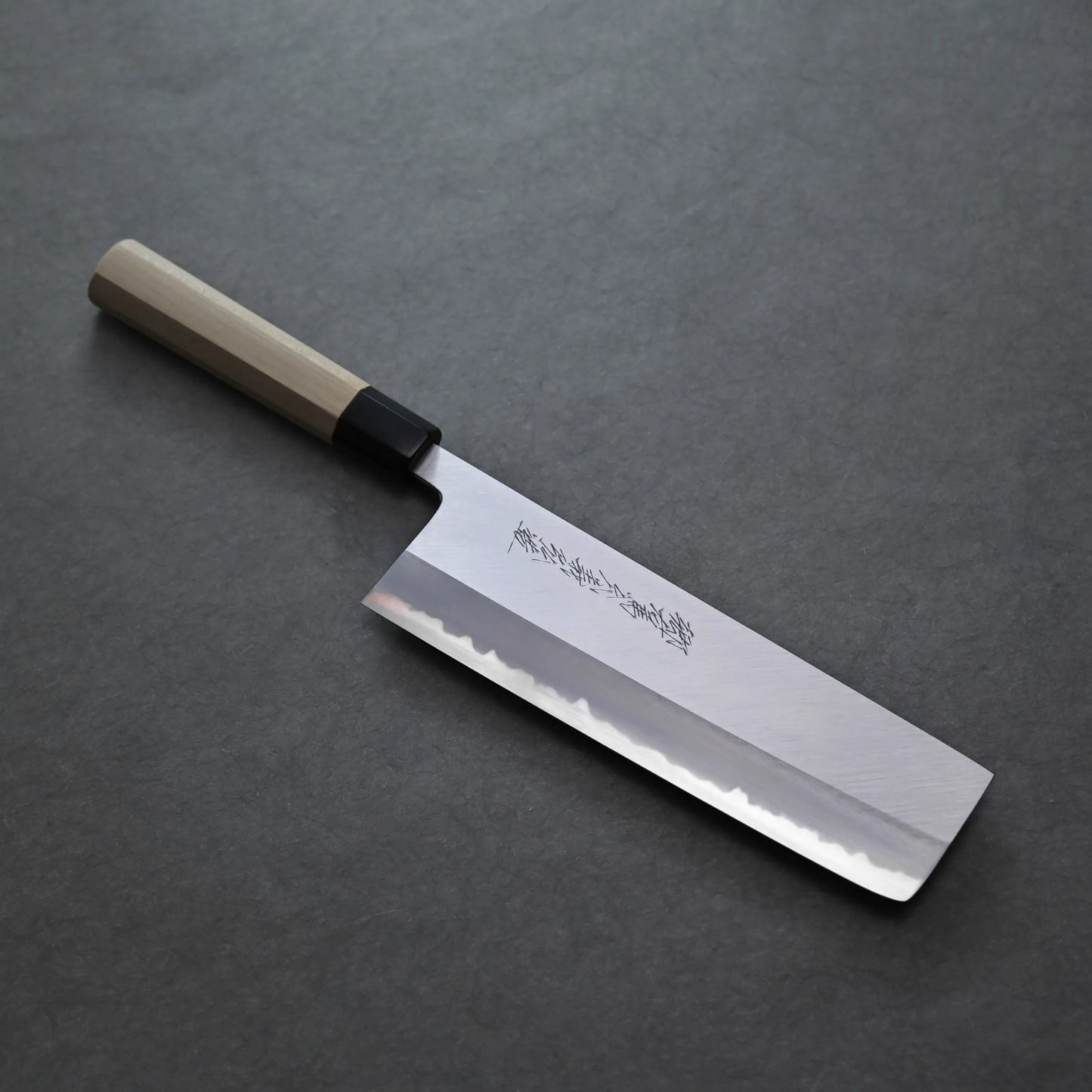 A Tanaka AS nakiri Japanese-style knife with a rectangular blade, wooden handle, and kanji characters etched on the blade, placed on a gray surface.