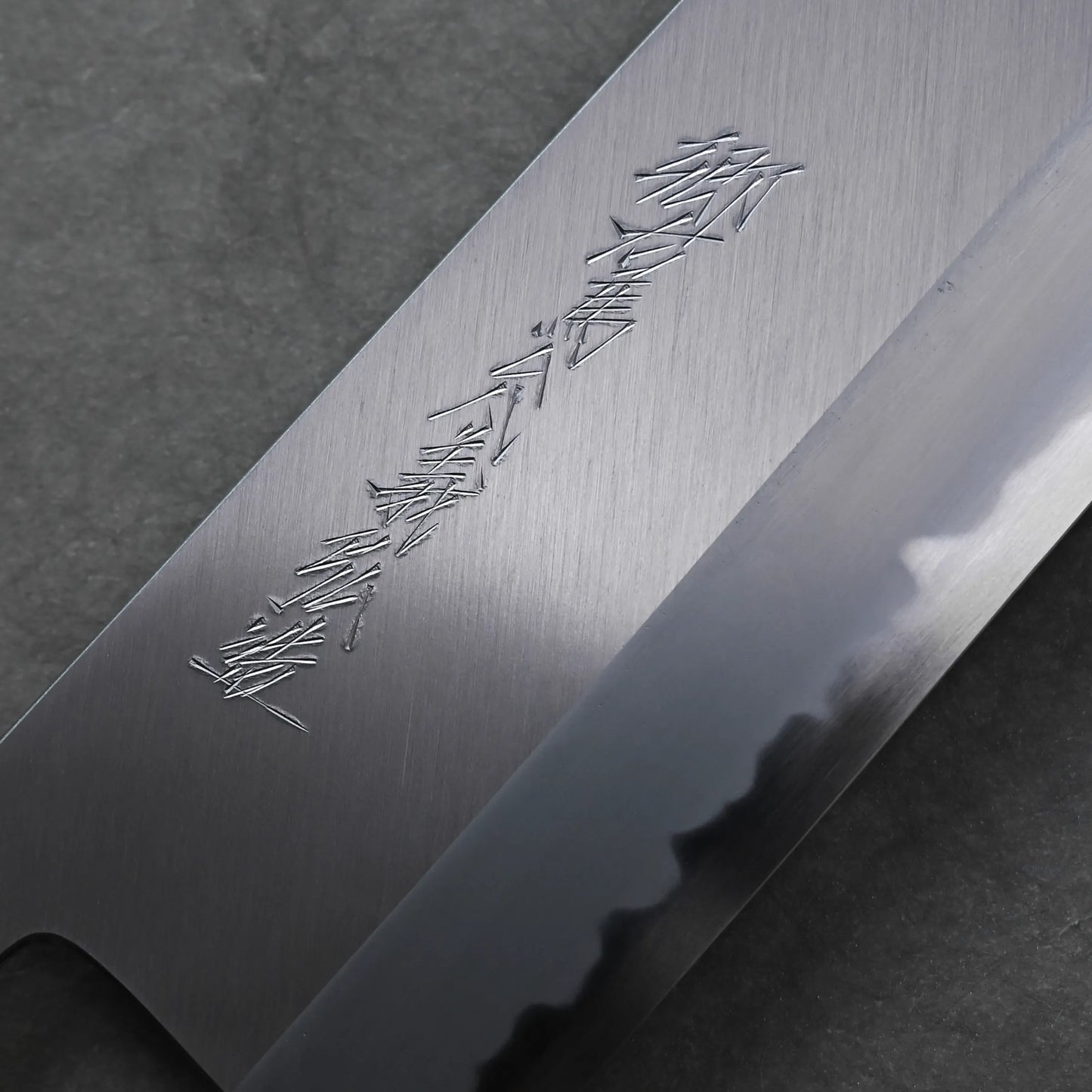 Close-up of a Tanaka 210mm nakiri blade with kanji characters engraved on the polished metal, displayed against a dark background.