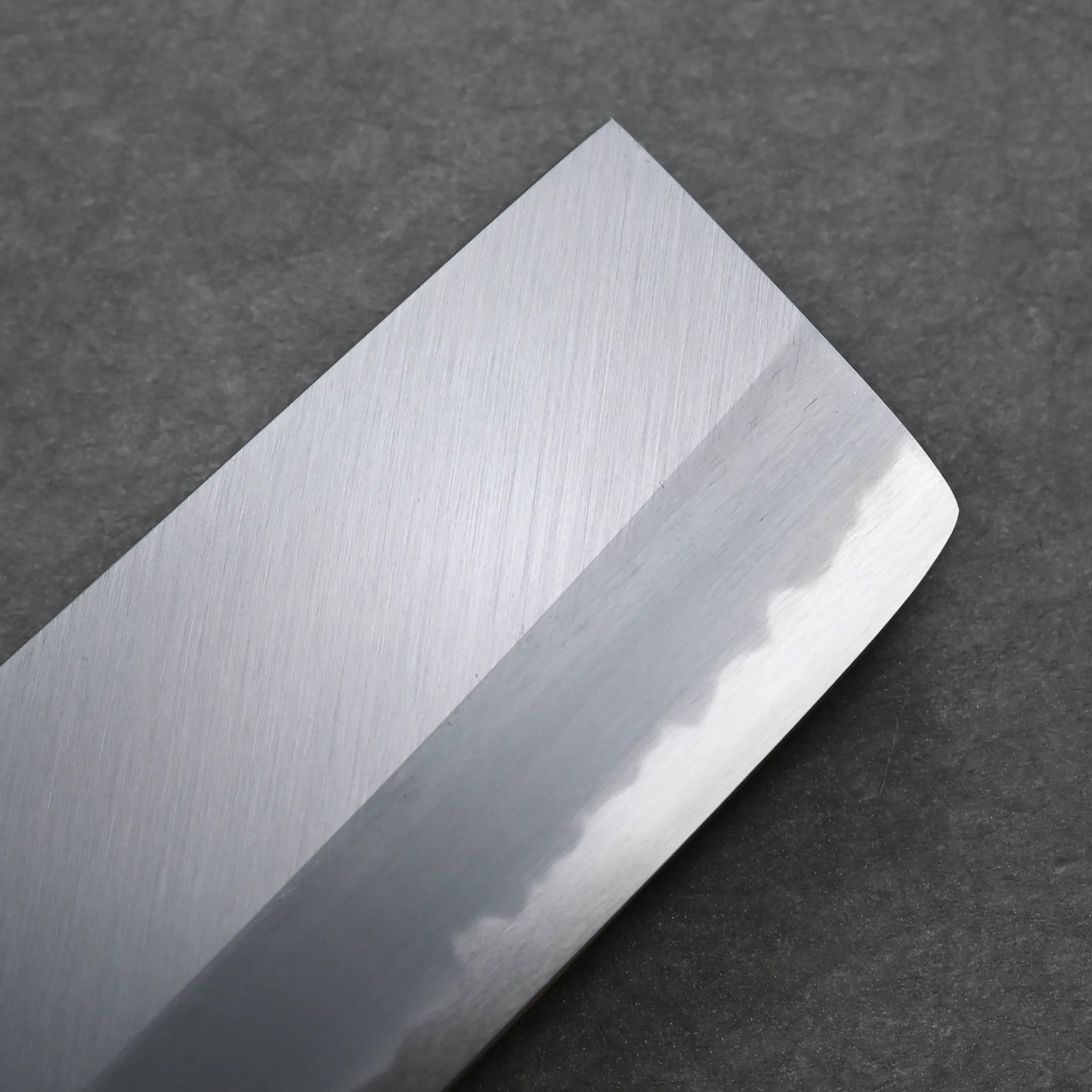 Close-up of a Tanaka AS nakiri blade with a straight edge, visible hamon line, and a slightly angled tip, set against a dark gray textured background.