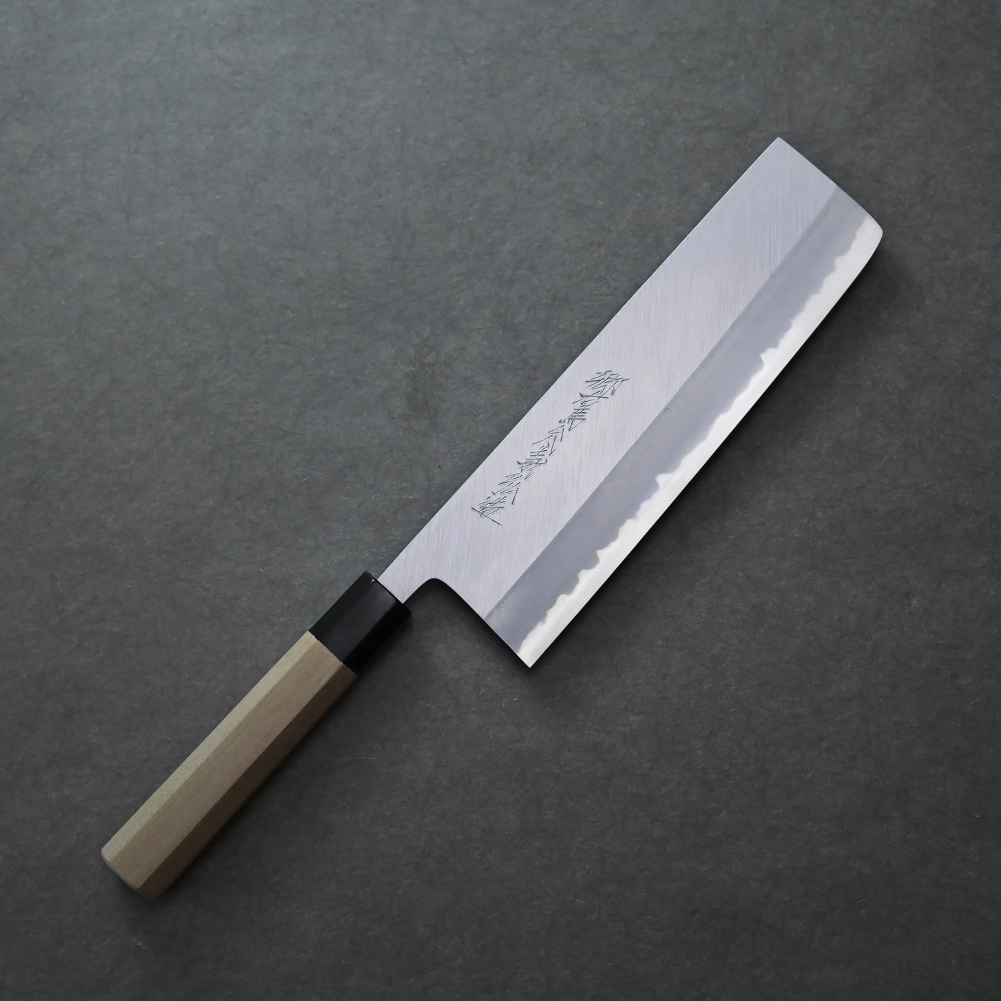 Yoshikazu Tanaka shinogi AS nakiri 210mm with a straight-edged blade and wooden handle rests on a dark gray surface. The blade features engraved characters and a polished finish.