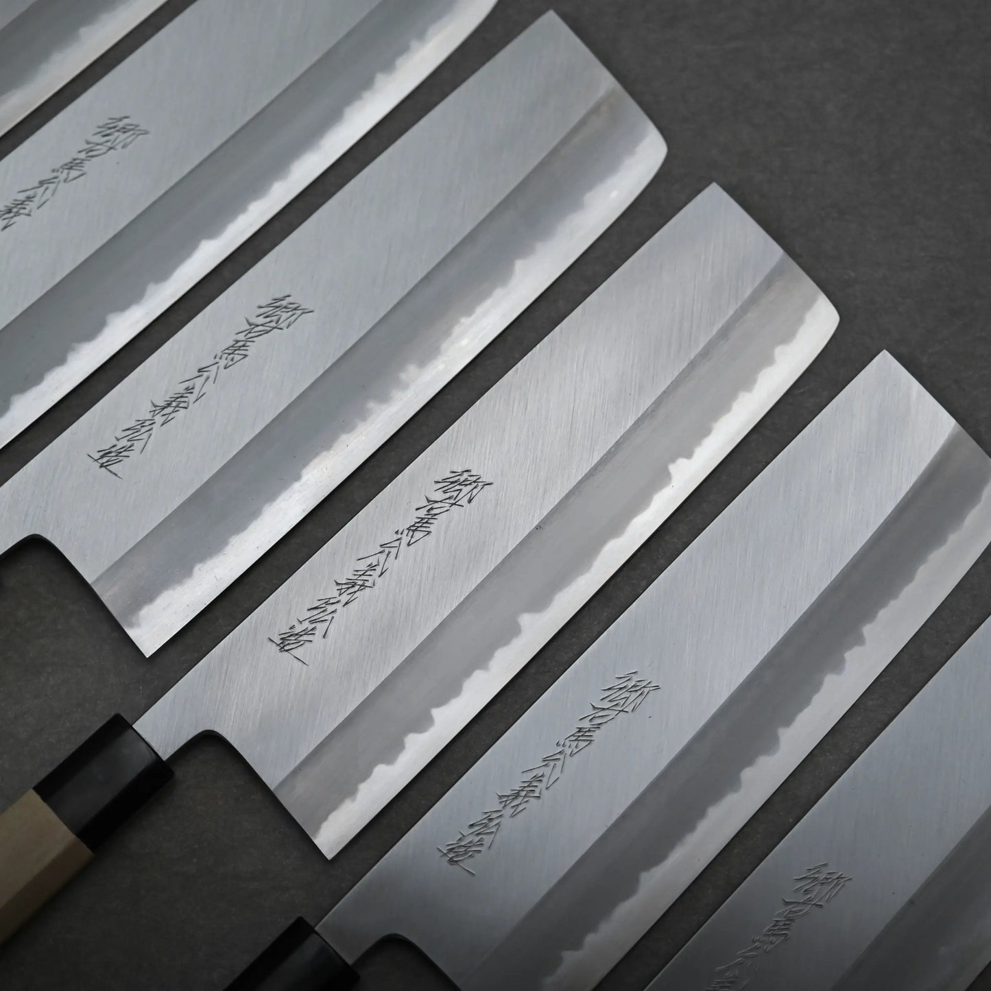 Set of four nakiri knives with engraved designs on their blades, arranged in a row on a dark surface.