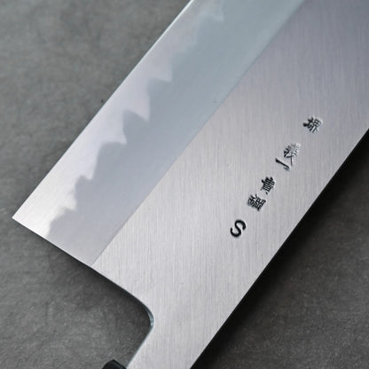 A close-up of a Tanaka AS nakiri blade, showing a wavy hamon line and engraved Japanese characters on the polished metal, resting on a dark, textured surface.