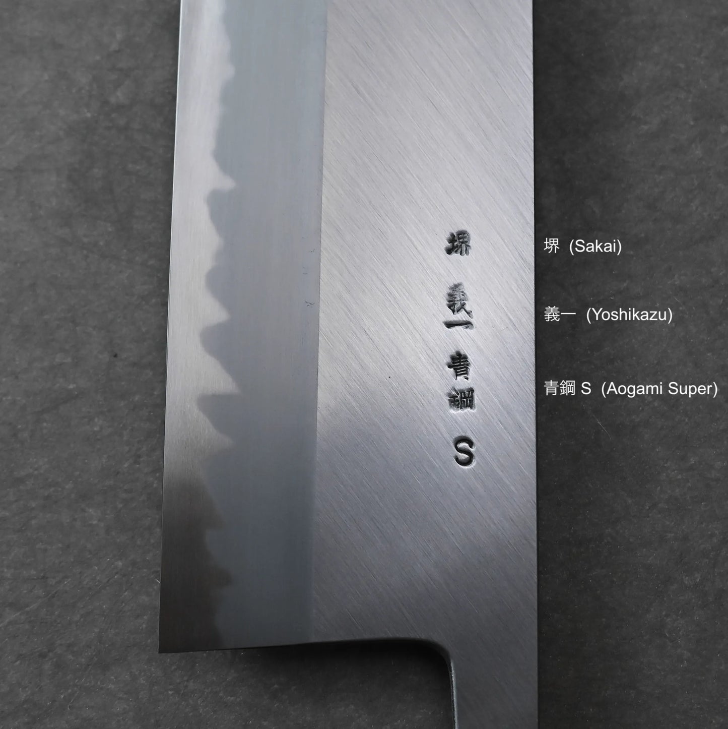 Close-up of a Japanese Tanaka AS nakiri blade with engraved kanji, placed on a dark surface. Text beside the blade translates the engravings as Sakai, Yoshikazu, and Aogami Super, with English labels in parentheses.