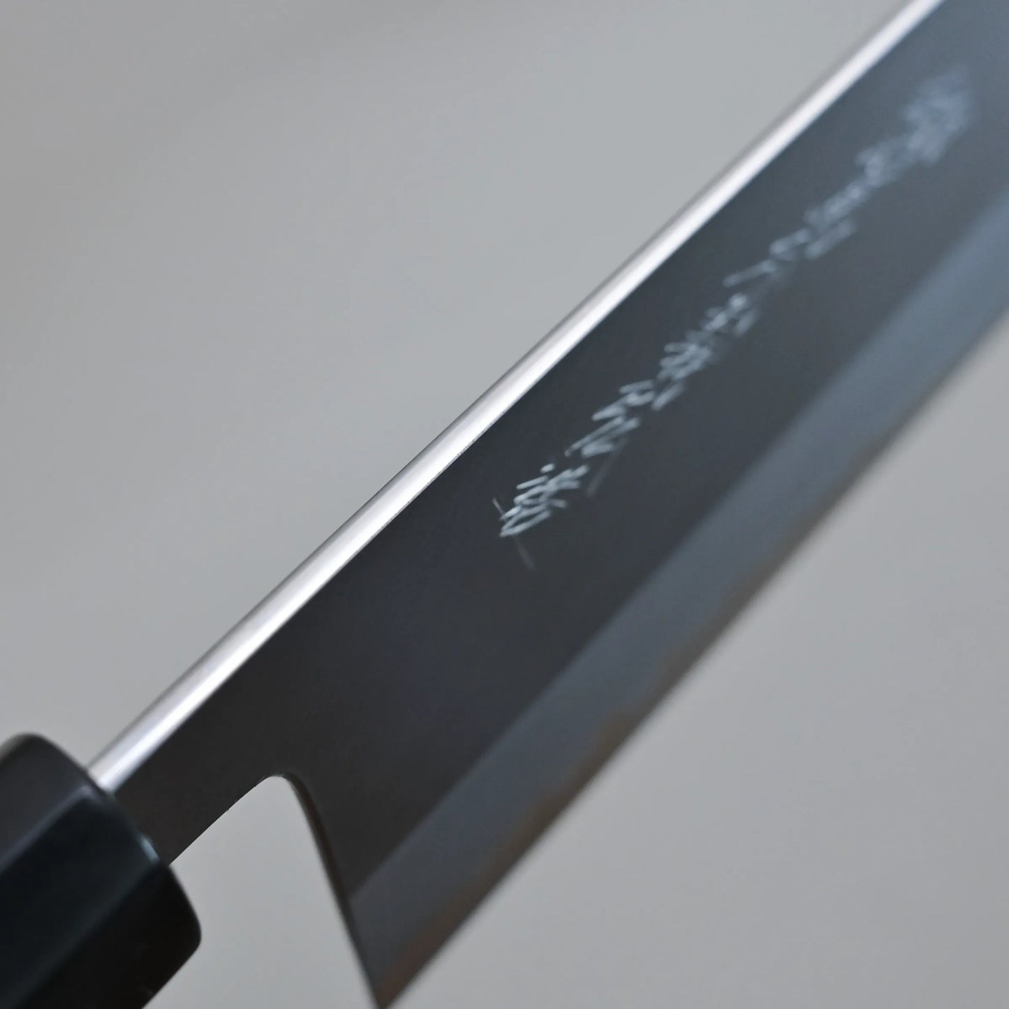 Close-up of the spine finish of a Tanaka 210mm nakiri blade with Japanese characters engraved on the polished metal. The background is out of focus, highlighting the fine detail and craftsmanship.