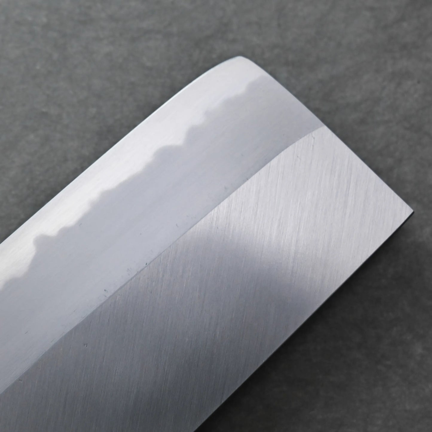 Close-up of a polished Tanaka AS nakiri blade with a visible wavy hamon line, set against a dark textured background.