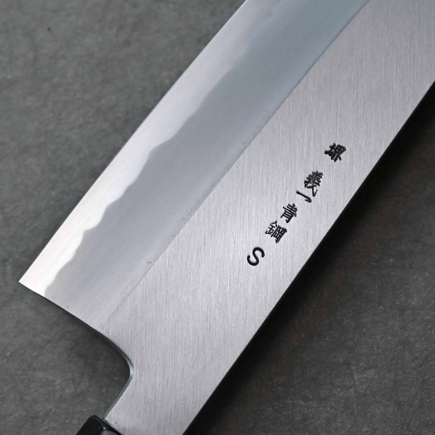 A close-up of the blade of a Tanaka AS gyuto shows a polished finish, wavy hamon line, and Japanese characters engraved on the metal. The background is a dark, textured surface.