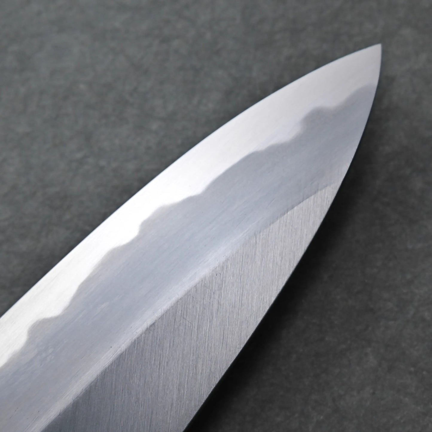 Close-up of a shiny Tanaka AS gyuto blade with a visible hamon pattern along the edge, set against a dark, textured background.