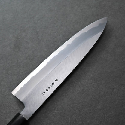 A close-up view of a Tanaka AS gyuto Japanese chef’s knife with a polished blade and kanji characters engraved near the handle, lying on a dark gray textured surface.