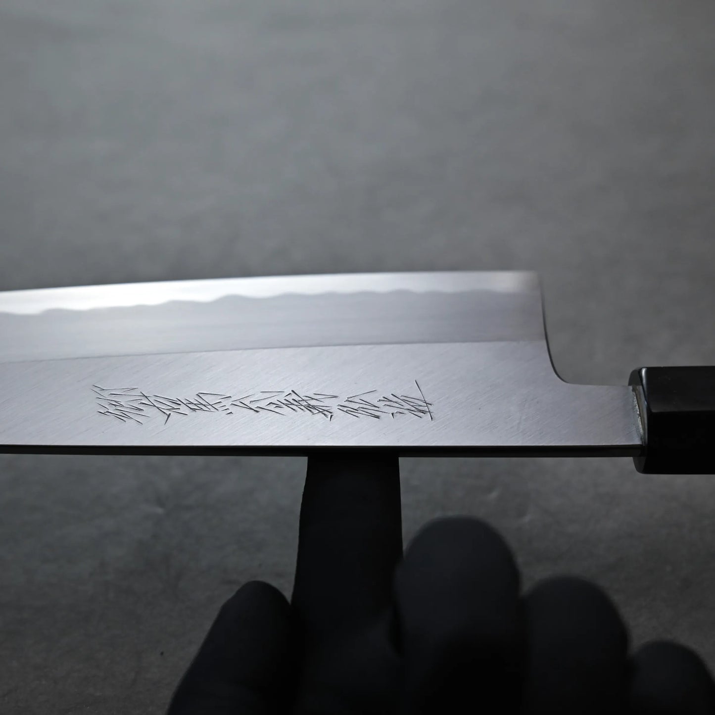 Balance point of  Tanaka AS gyuto carbon steel knife blade with engraved markings, held by a person wearing a black glove, against a plain gray background.