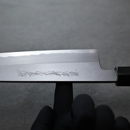 Balance point of  Tanaka AS gyuto carbon steel knife blade with engraved markings, held by a person wearing a black glove, against a plain gray background.