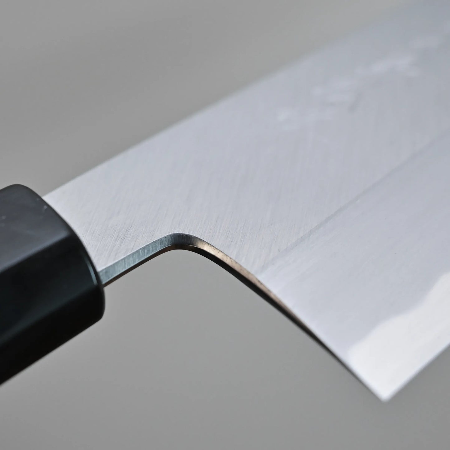Close-up view of choil of a Tanaka AS gyuto, set against a neutral grey background. The clean surface of the carbon steel blade is clearly visible.