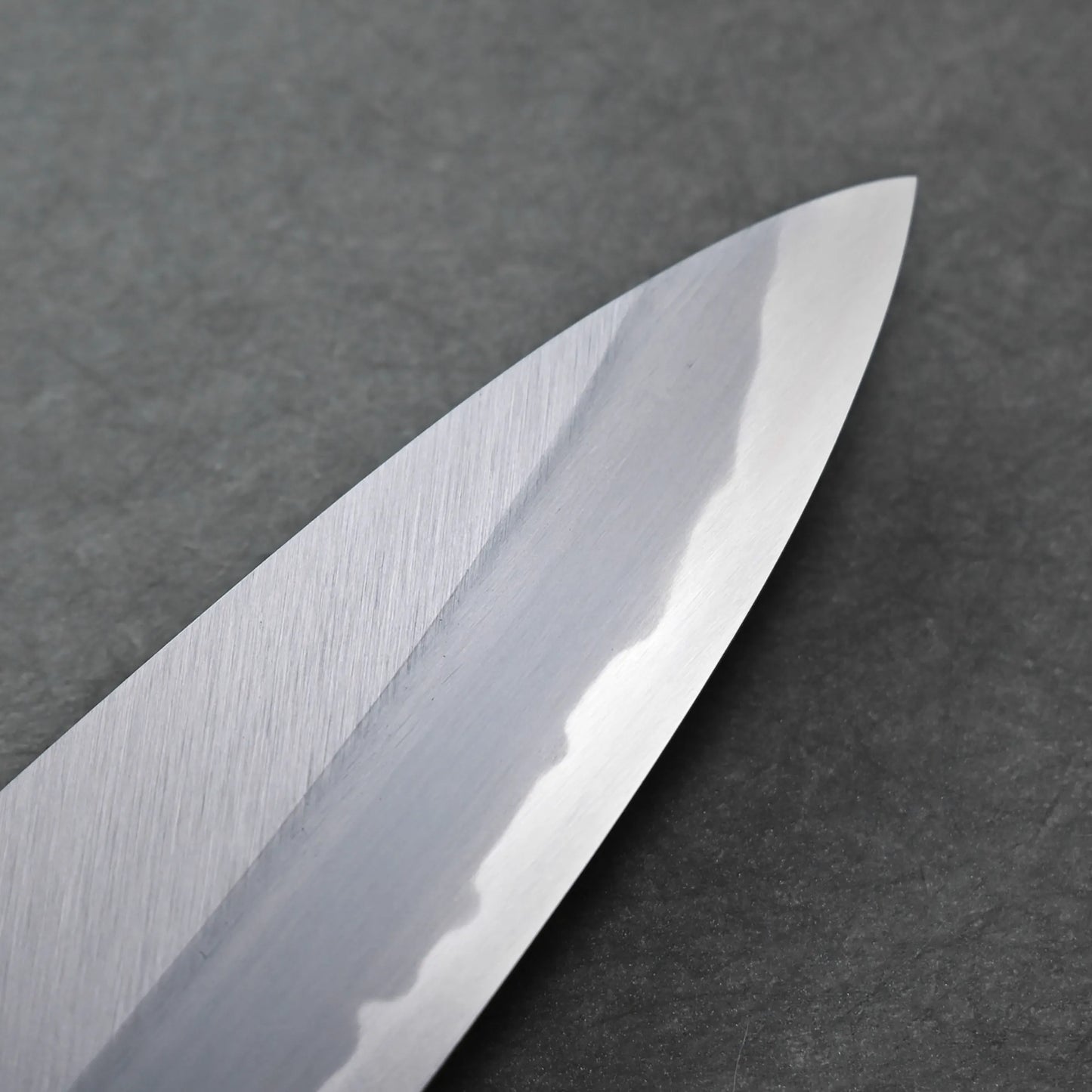 Close-up of a polished Tanaka AS tall gyuto blade with a visible wavy hamon line, placed on a dark grey textured surface.