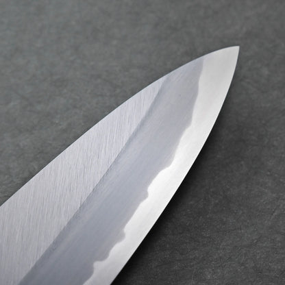 Close-up of a polished Tanaka AS tall gyuto blade with a visible wavy hamon line, placed on a dark grey textured surface.