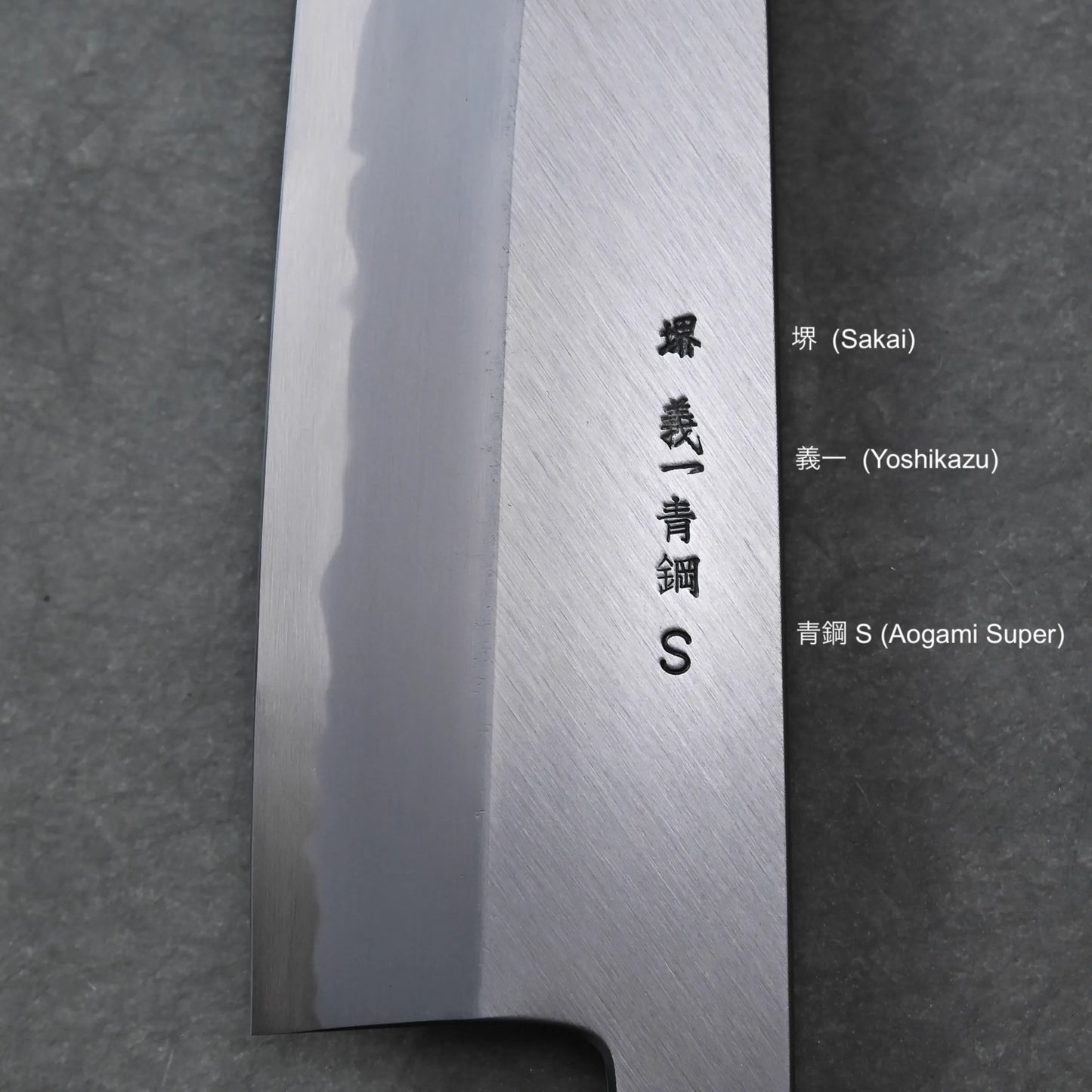 Close-up of a Tanaka AS gyuto blade with engraved kanji characters and the letter S, along with English translations—Sakai, Yoshikazu, and Aogami Super—set against a dark textured background.