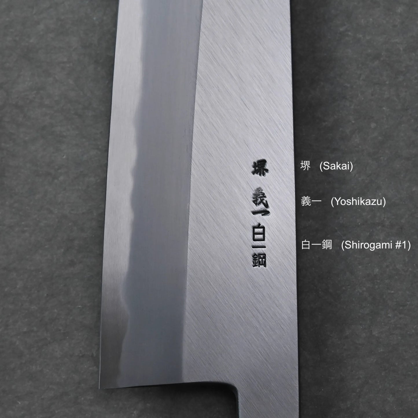 A close-up of a Japanese knife blade with engraved kanji characters and a polished edge. Text on the right provides translations: Sakai, Yoshikazu, and Shirogami #1 (White Steel). The background is dark gray.