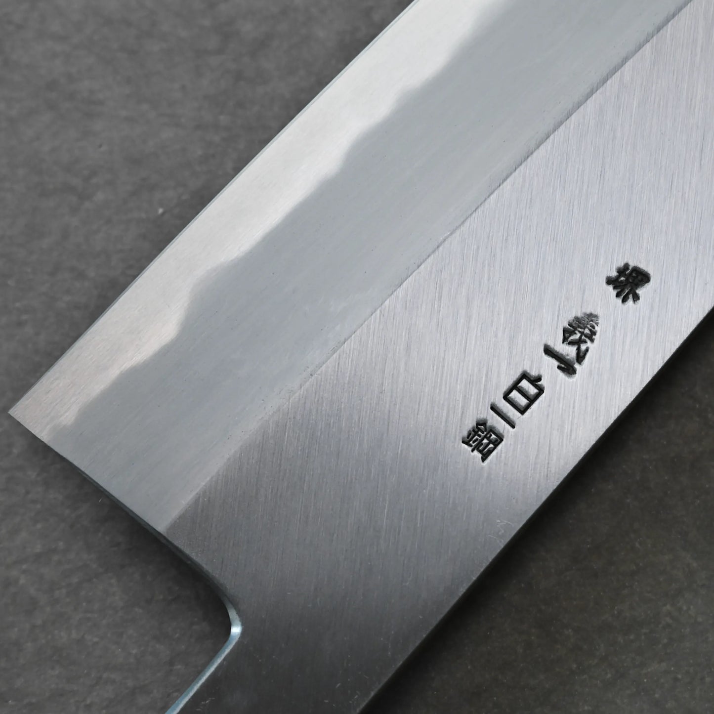 Close-up of a Japanese knife blade with a visible hamon line and engraved kanji characters, displayed on a dark, textured surface.