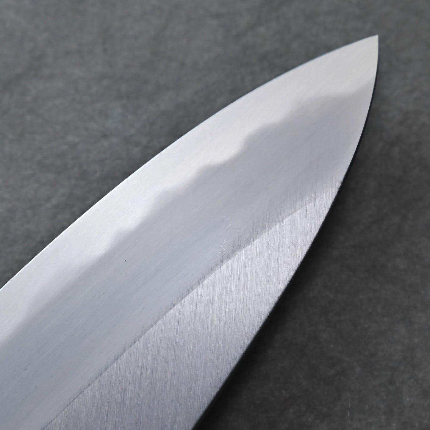Close-up of a shiny, silver knife blade featuring a visible wavy pattern along the edge against a dark, textured background, highlighting its elegant craftsmanship.