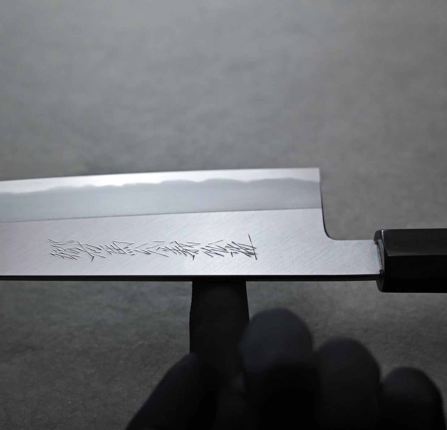 Balance point of Tanaka tall gyuto with Japanese characters engraved on the blade. A gloved finger points to the engraving, while the gray background remains out of focus.