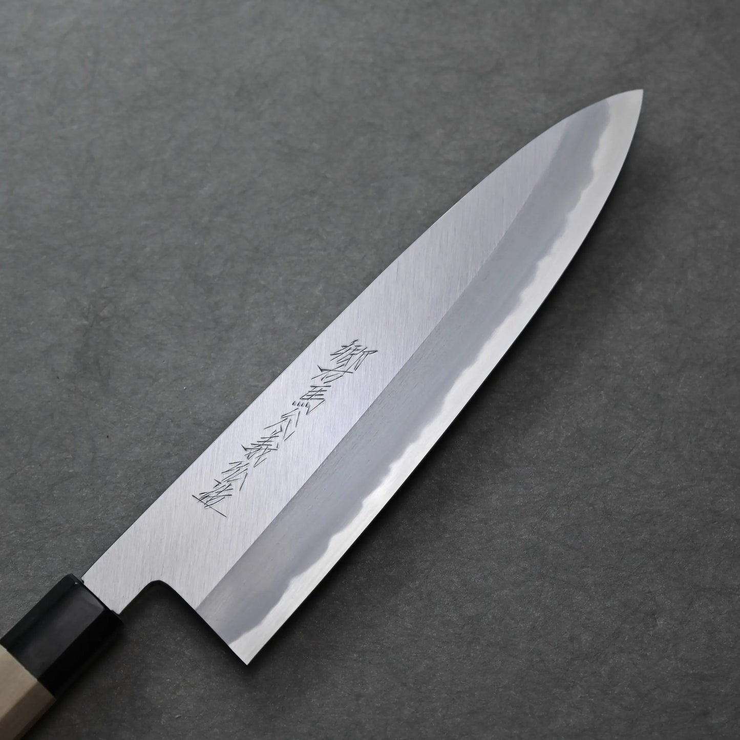 A close-up of a Japanese-style knife with a polished steel blade, visible hamon line, engraved characters, and a wooden handle resting on a dark grey textured surface.