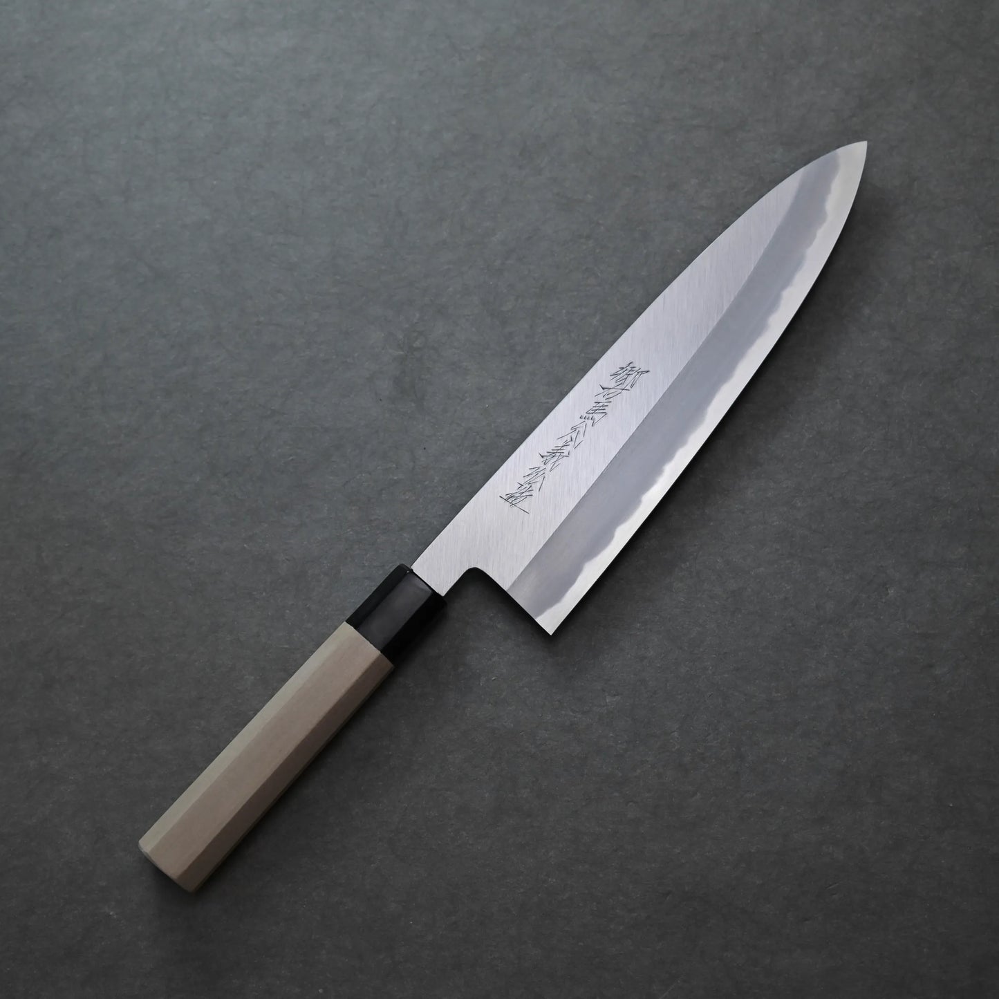 Yoshikazu Tanaka shinogi shirogami#1 tall gyuto 240mm with a wooden handle and a polished, engraved blade rests on a dark, textured surface, showcasing exquisite craftsmanship.