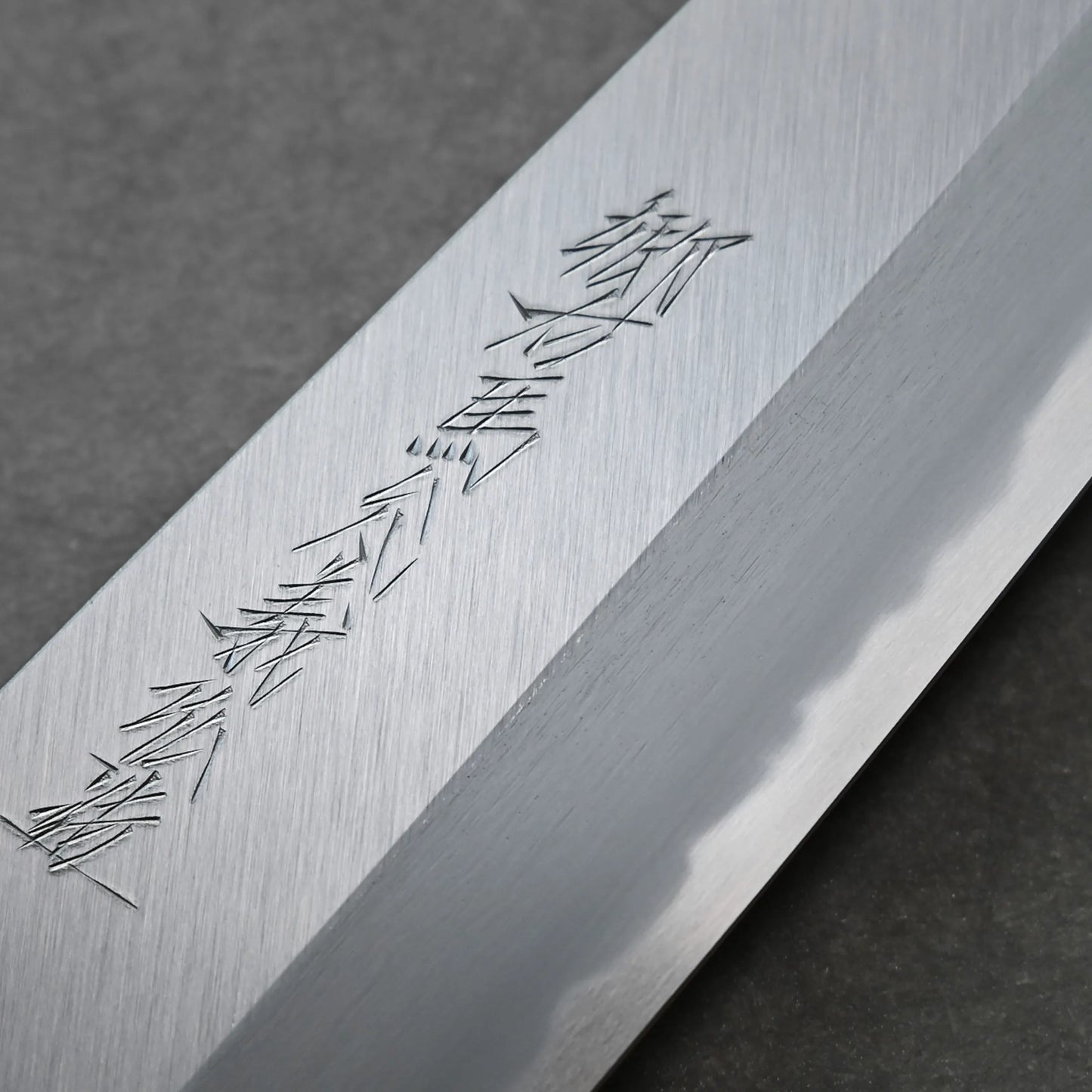 Close-up of a polished knife blade featuring Japanese kanji characters engraved on the surface, displayed against a dark, textured background for an authentic aesthetic.