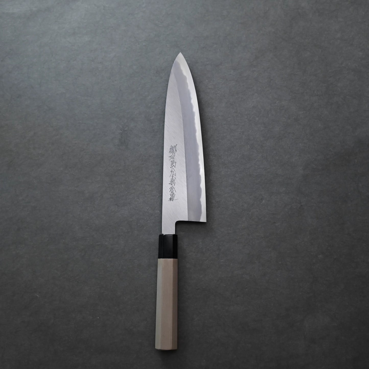 A Japanese-style knife with a wooden handle and a gleaming, slightly curved blade engraved with Japanese characters rests on a gray surface.